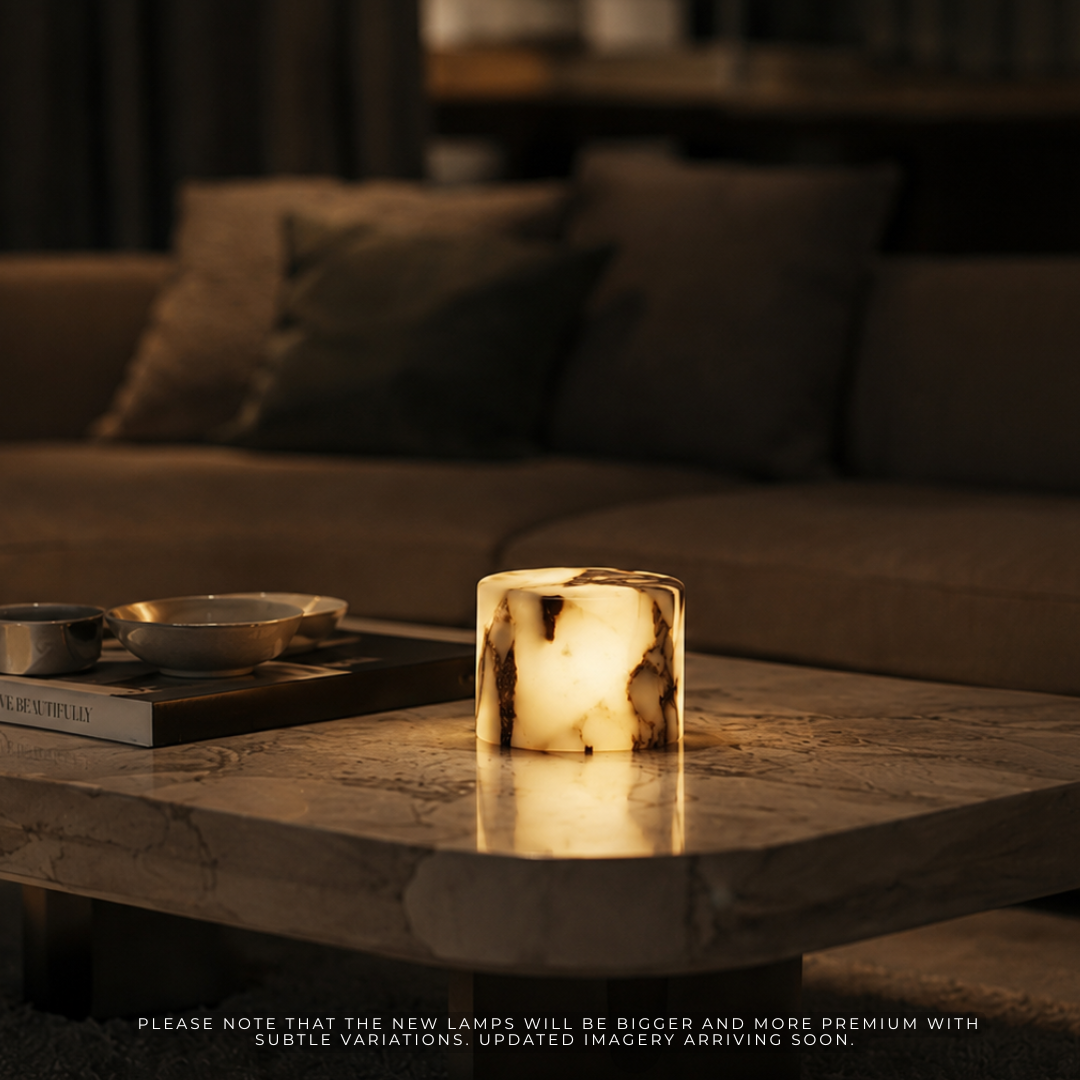 A cylindrical table lamp with a glowing marbled stone or onyx-like finish sits on a marble coffee table in a dimly lit living room, with a sofa and books blurred in the background.