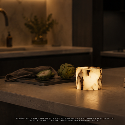 A cylindrical lamp with a white and black marbled stone-like exterior glowing warm yellow inside, placed on a kitchen countertop in a dimly lit modern kitchen setting.