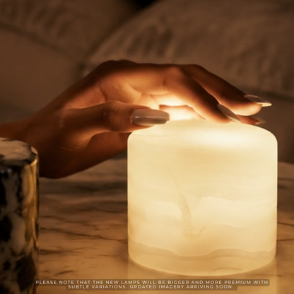 A warm white LED night light shaped like a stylized tree, glowing softly on a marble surface with a hand resting above it. The scene is warmly lit, and a small portion of a patterned mug is visible on the left.