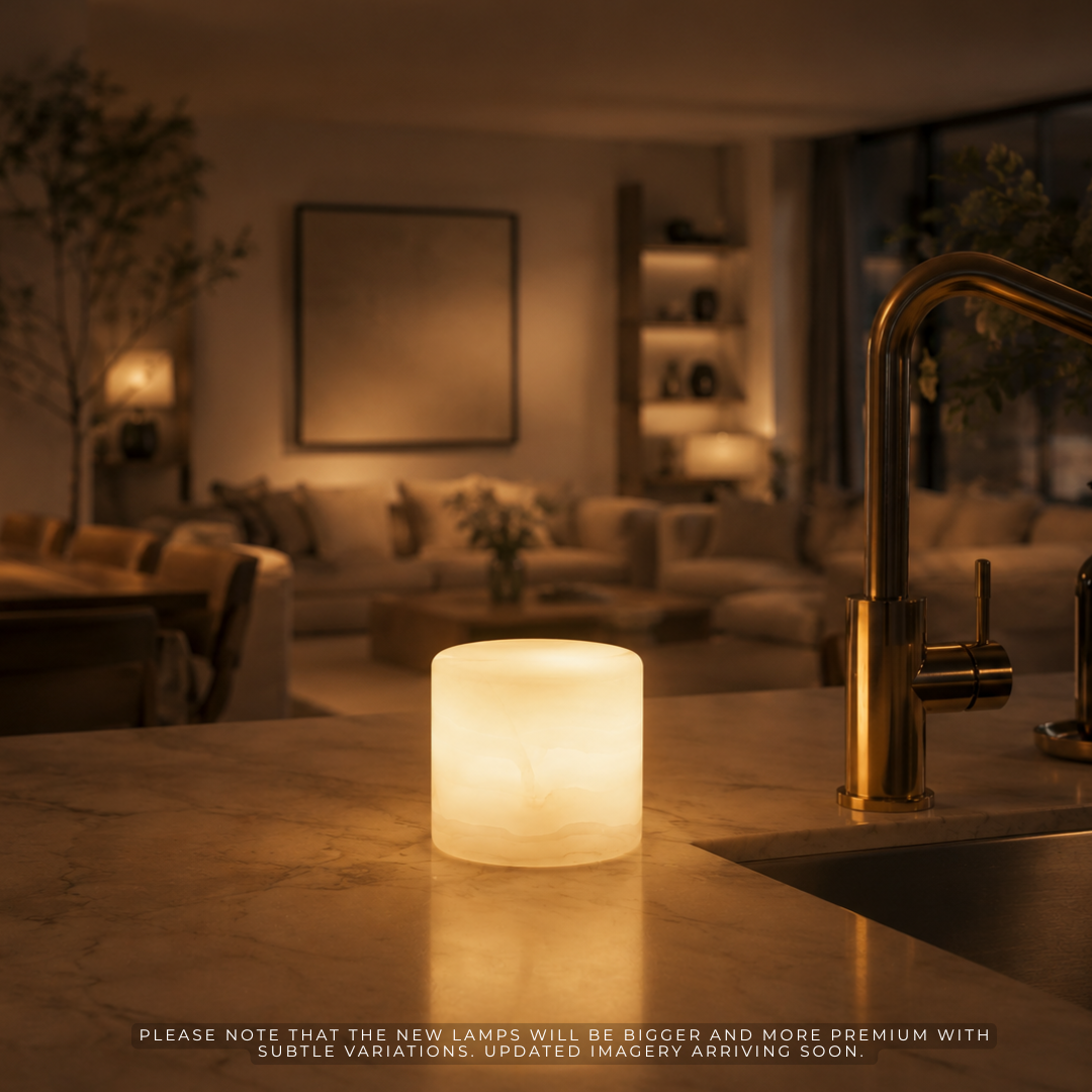 A glowing cylindrical lamp sits on a kitchen or bathroom countertop near a brass faucet, with a softly blurred living room in the background. The lamp emits a warm white light, and small disclaimer text appears along the bottom edge.